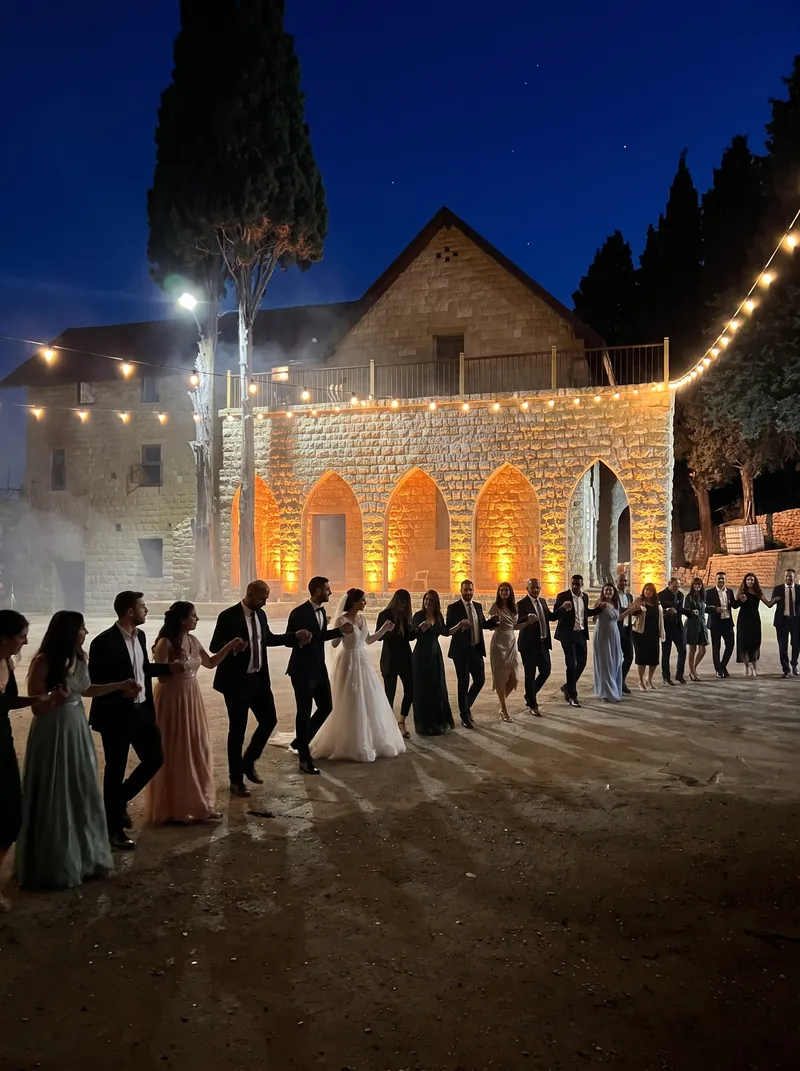 A bride, groom and guests dancing dabke in a long line, holding hands, in front of the lit arches of Qasr Noun at night under festoon lights, shown as a styled wedding visualisation
