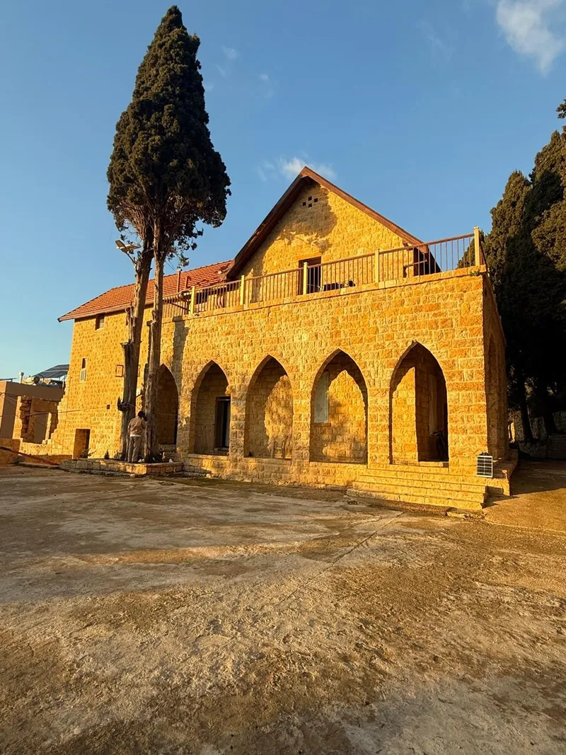 The restored stone arcade of Qasr Noun in Ain Anub, five pointed arches under a tiled roof, beside a tall cypress at golden hour