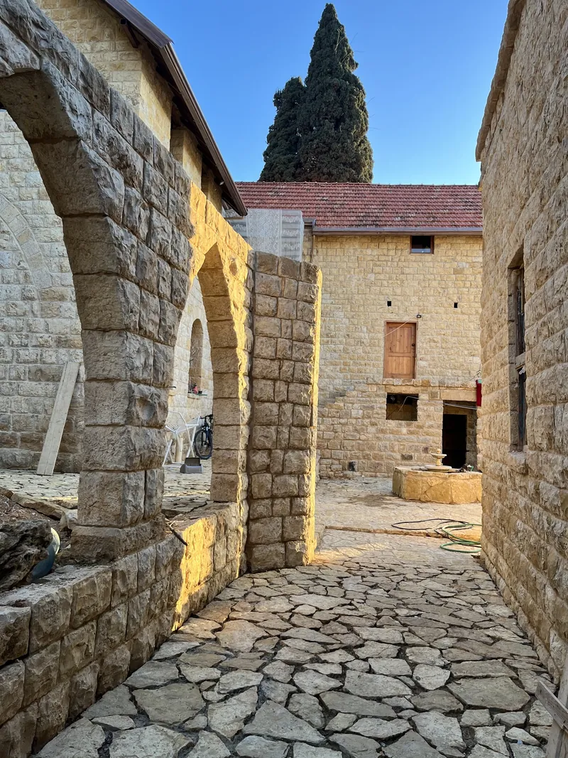 A paved stone lane at Qasr Noun between restored stone walls and an arch, with a stone house and a tall cypress beyond, in daylight