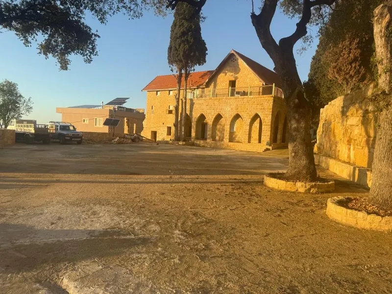 The stone house and arched arcade of Qasr Noun in Ain Anub, seen across the courtyard under old trees in late-afternoon sun